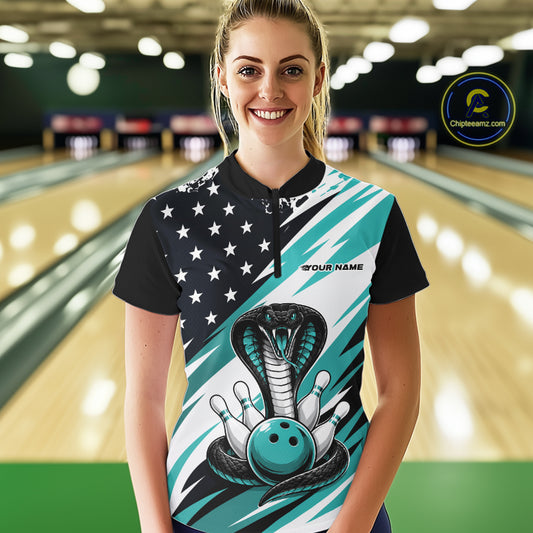 A woman stands in a bowling alley wearing a black, white, and blue bowling shirt with stars, diagonal stripes, a cobra, pins, and an blue ball.