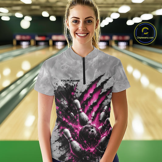 Front view of a female model wearing a gray bowling quarter zip shirt featuring a dramatic pink bowling ball and pins graphic with claw-mark artwork at a bowling lane.