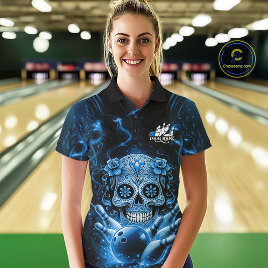 Female model at a bowling lane wearing a black and blue polo shirt with sugar skull artwork and bowling strike-themed graphics.