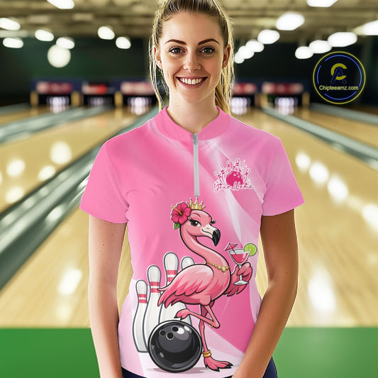Female model wearing a pink bowling quarter-zip shirt at a bowling lane with a crowned flamingo, cocktail, bowling ball, and hibiscus flower accents.