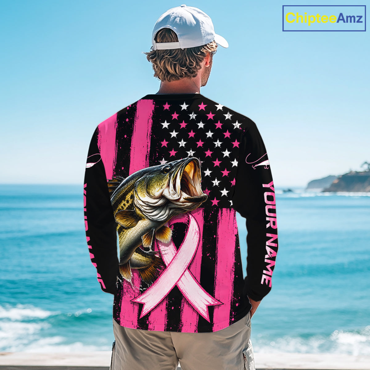 A man viewed from behind wearing a long-sleeve black and pink graphic shirt with a large fish, stars, stripes, and a pink ribbon on the back, standing by the ocean. The shirt has 'YOUR NAME' printed vertically on the sleeve.