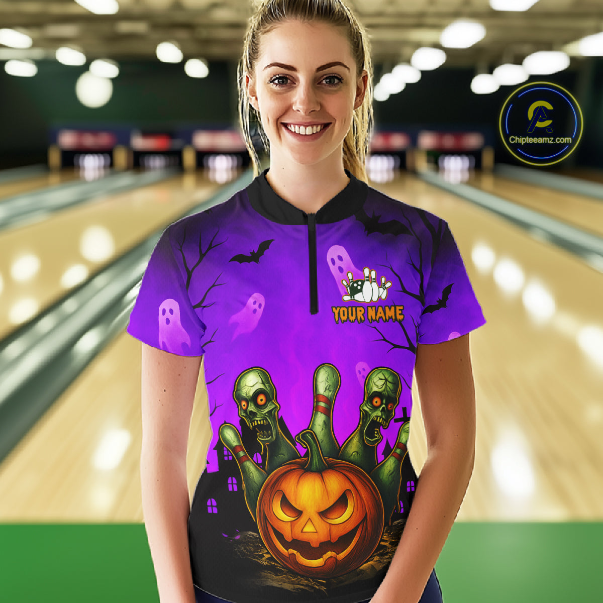 A woman stands in a bowling alley wearing a short-sleeve purple Halloween-themed polo shirt with a black collar and a full-front graphic featuring a jack-o'-lantern, green skeletons, ghosts, bats, and the text 'YOUR NAME'.