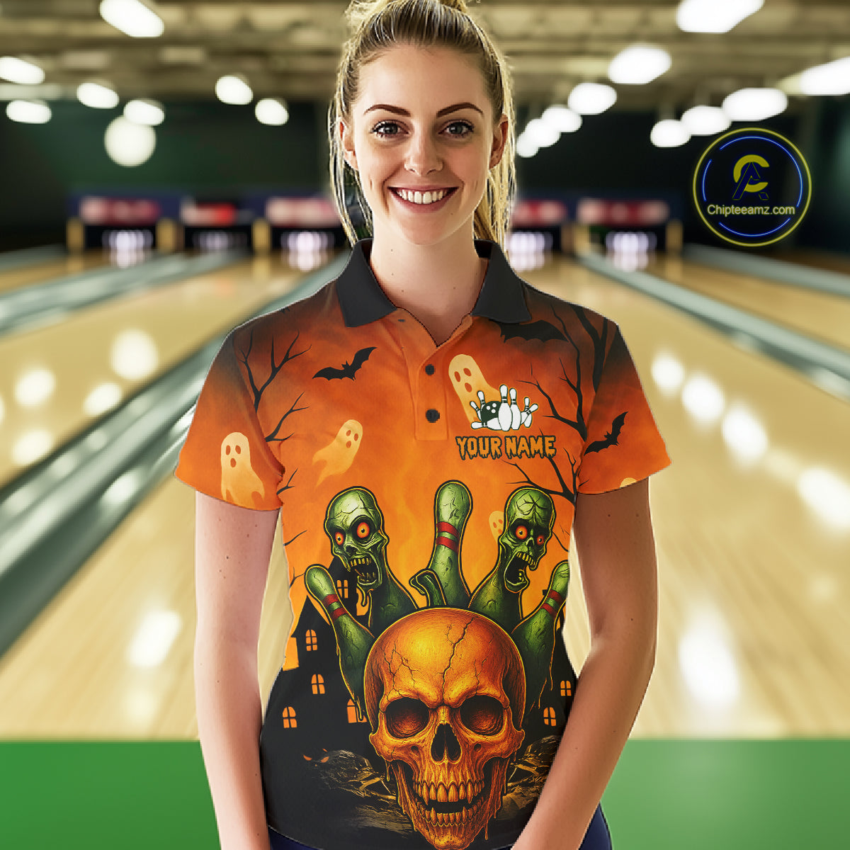 A woman stands in a bowling alley wearing a short-sleeve polo shirt with a Halloween-themed graphic print featuring a skull, green zombies, ghosts, and bats on an orange background. The shirt has a dark collar and the text 'YOUR NAME' on the chest.