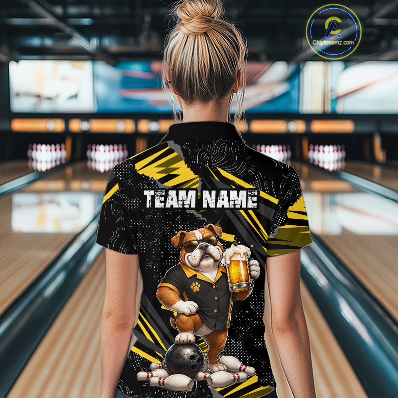 A woman viewed from behind wearing a black and yellow bowling-themed short-sleeve polo shirt with a bulldog graphic holding a beer mug and the text 'TEAM NAME'. The background shows a blurred bowling alley with lanes and pins.