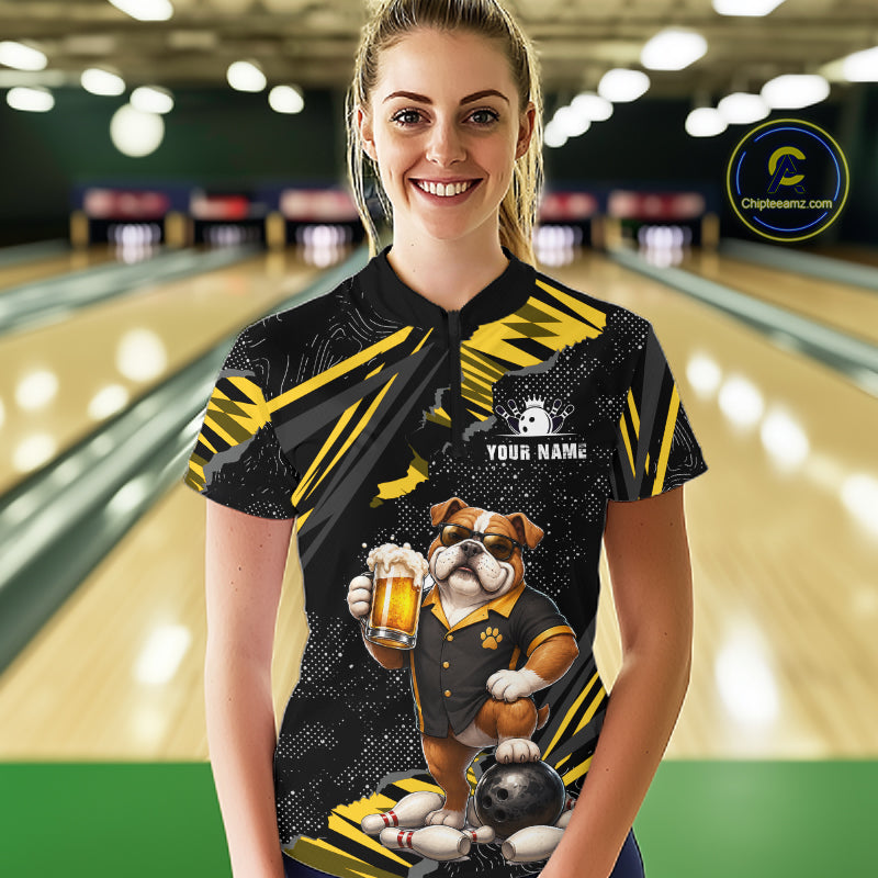 A woman standing in a bowling alley wearing a custom black and yellow short-sleeve bowling shirt with a bulldog graphic holding a beer mug and a bowling ball. The shirt has a custom name placeholder on the chest.