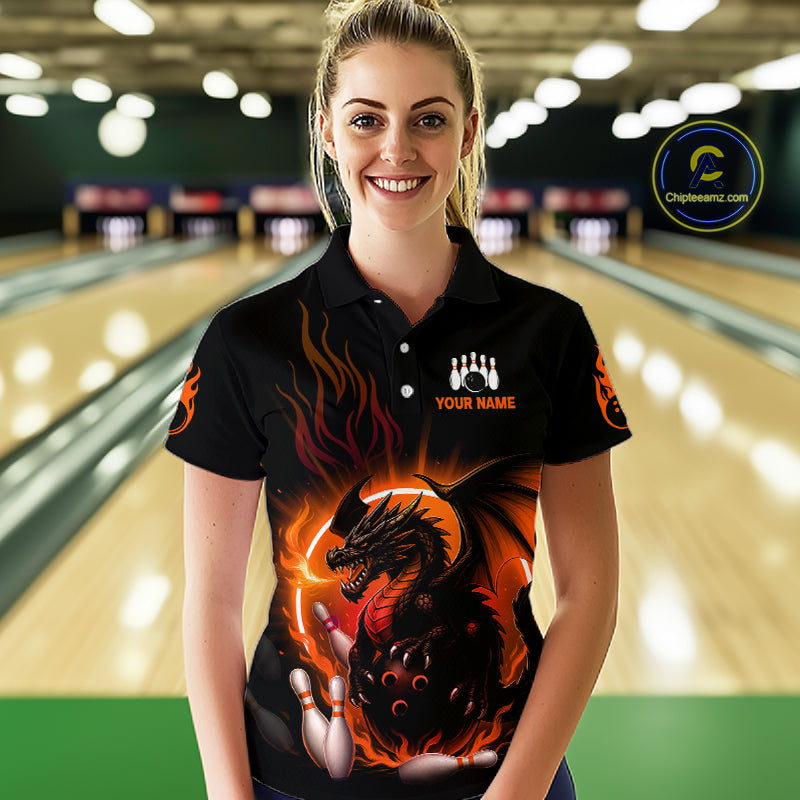 A woman wearing a black short-sleeve bowling polo shirt with a fiery dragon graphic and white text, standing in a bowling alley with lanes blurred in the background.