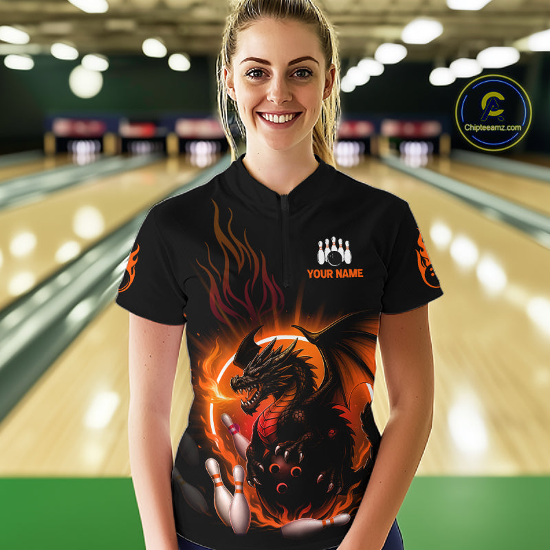A woman stands in a bowling alley wearing a black short-sleeve bowling shirt with a large fiery dragon graphic on the front, orange flame details, and a white custom name patch on the chest. 