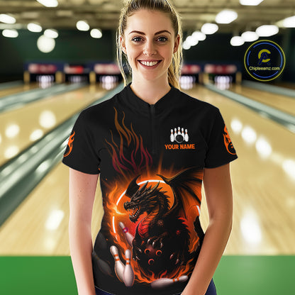 A woman stands in a bowling alley wearing a black short-sleeve bowling shirt with a large fiery dragon graphic on the front, orange flame details, and a white custom name patch on the chest. 