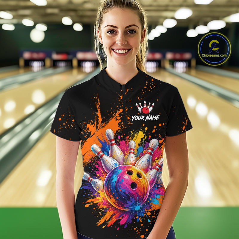 A woman wearing a black bowling-themed polo shirt with a colorful bowling ball and pins graphic, standing in a bowling alley. The shirt features a white crown logo, the text 'YOUR NAME', and a colorful paint-splatter design.