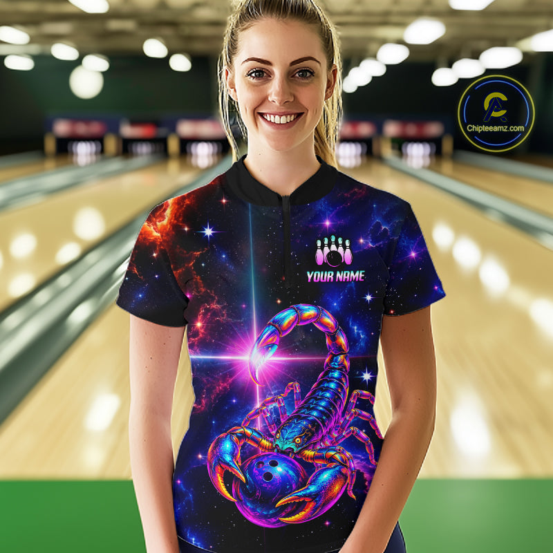 A woman stands in a bowling alley wearing a short-sleeve collared shirt with a vivid cosmic scorpion graphic on a black background. The shirt has a custom name printed on the chest