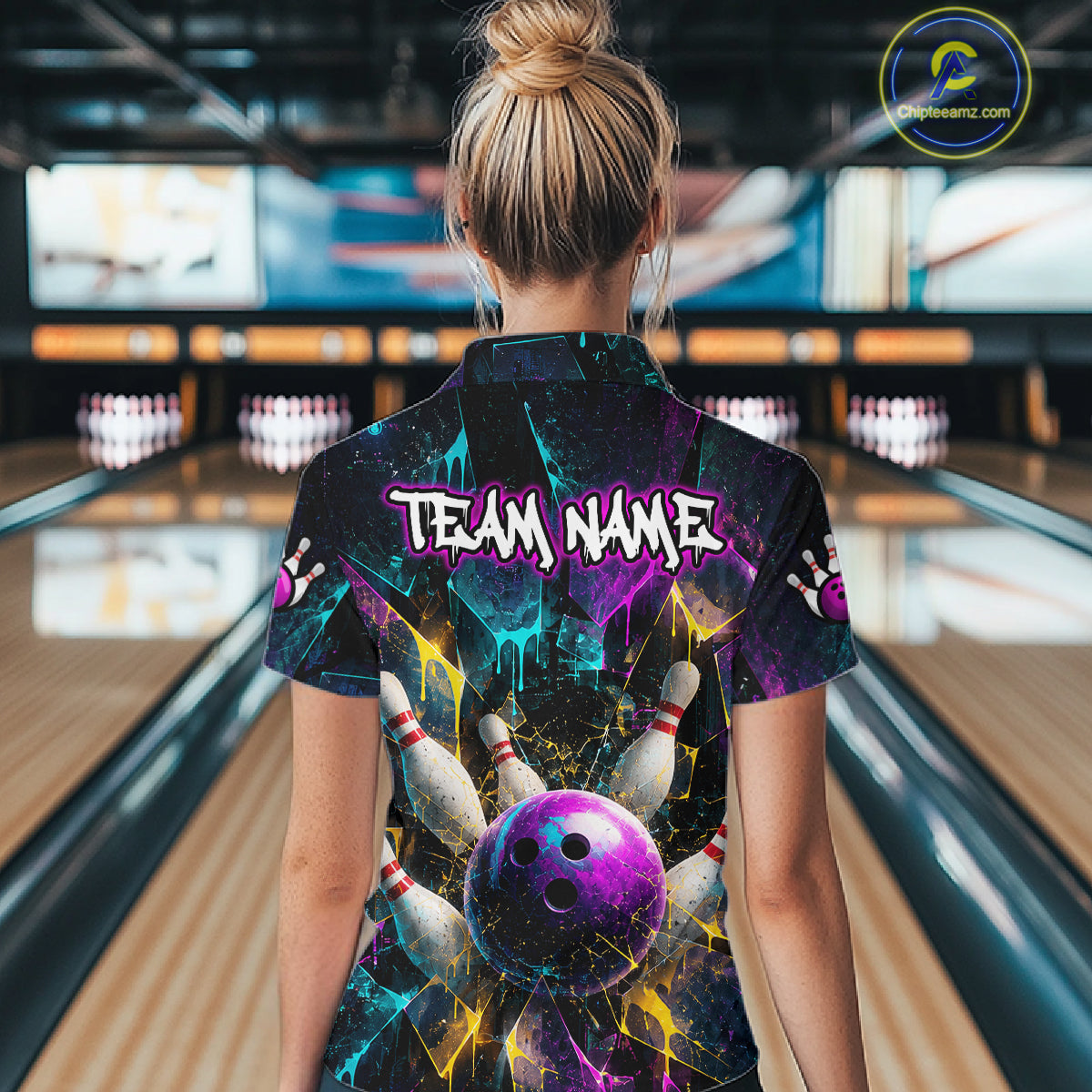 A woman viewed from behind wearing a short-sleeve bowling-themed shirt with a colorful bowling ball and pins graphic and the text 'TEAM NAME' on the back, standing in a bowling alley.