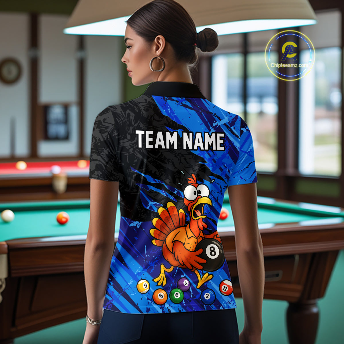 A woman stands in a poolroom wearing a short-sleeve collared shirt with a custom blue and black graphic design featuring a cartoon chicken, billiard balls, and the text 'TEAM NAME'.