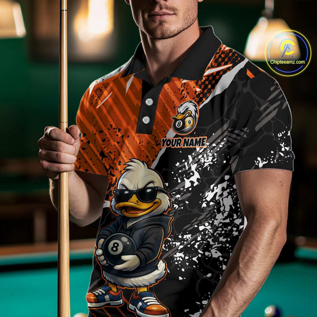 A man wearing a custom-designed short-sleeve polo shirt with a split orange and black pattern, featuring a cartoon duck character holding an 8-ball and the text 'YOUR NAME'. He is holding a pool cue in a dimly lit pool hall setting.