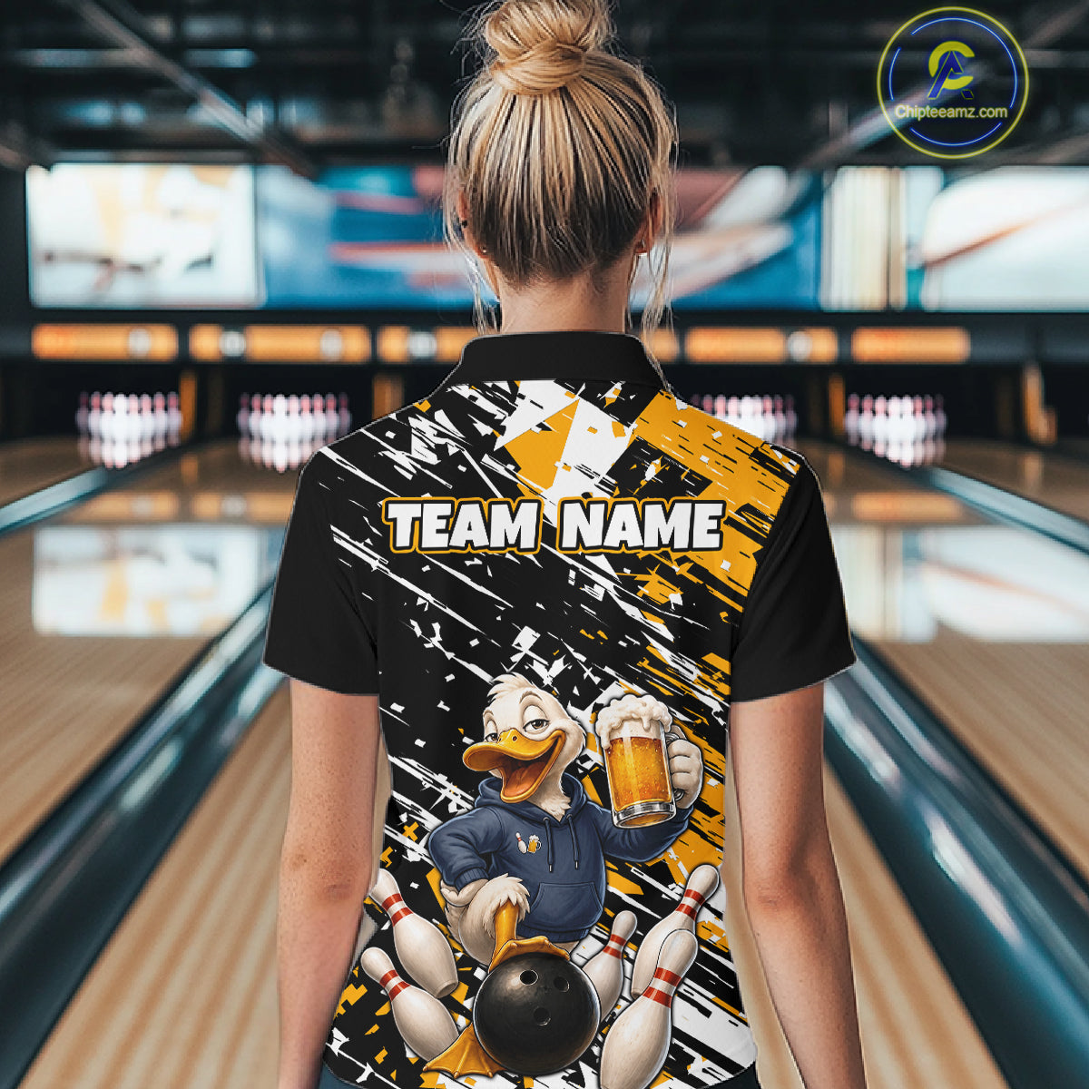 A woman viewed from behind wearing a black short-sleeve bowling-themed polo shirt with a large cartoon duck graphic, bowling pins, and the text 'TEAM NAME' on the back. The background shows a blurred bowling alley interior with lanes and pins.