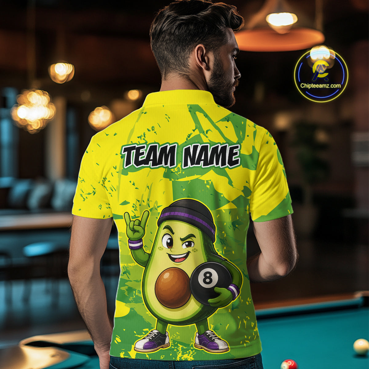 Person wearing a yellow and green shirt with an avocado character holding a pool ball, standing in a bar.
