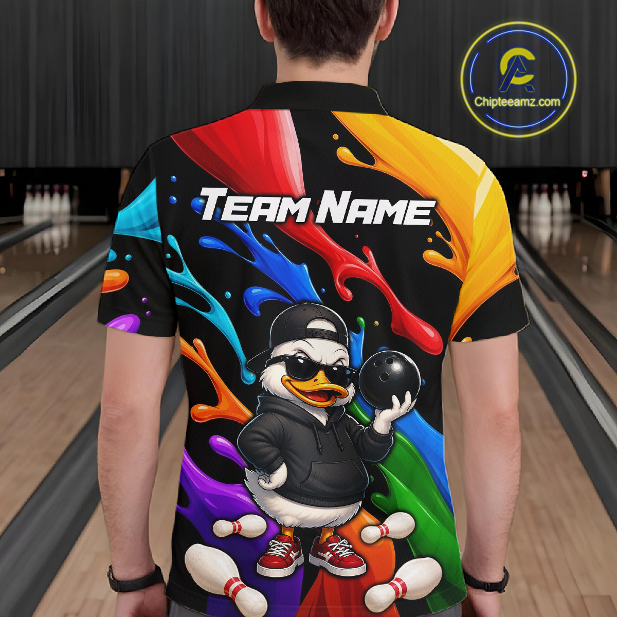 A person shown from behind wearing a custom short-sleeve bowling shirt with a colorful paint-splatter design, a cartoon duck character holding a bowling ball, and the text 'TEAM NAME' on the back. Bowling lanes are visible in the background.