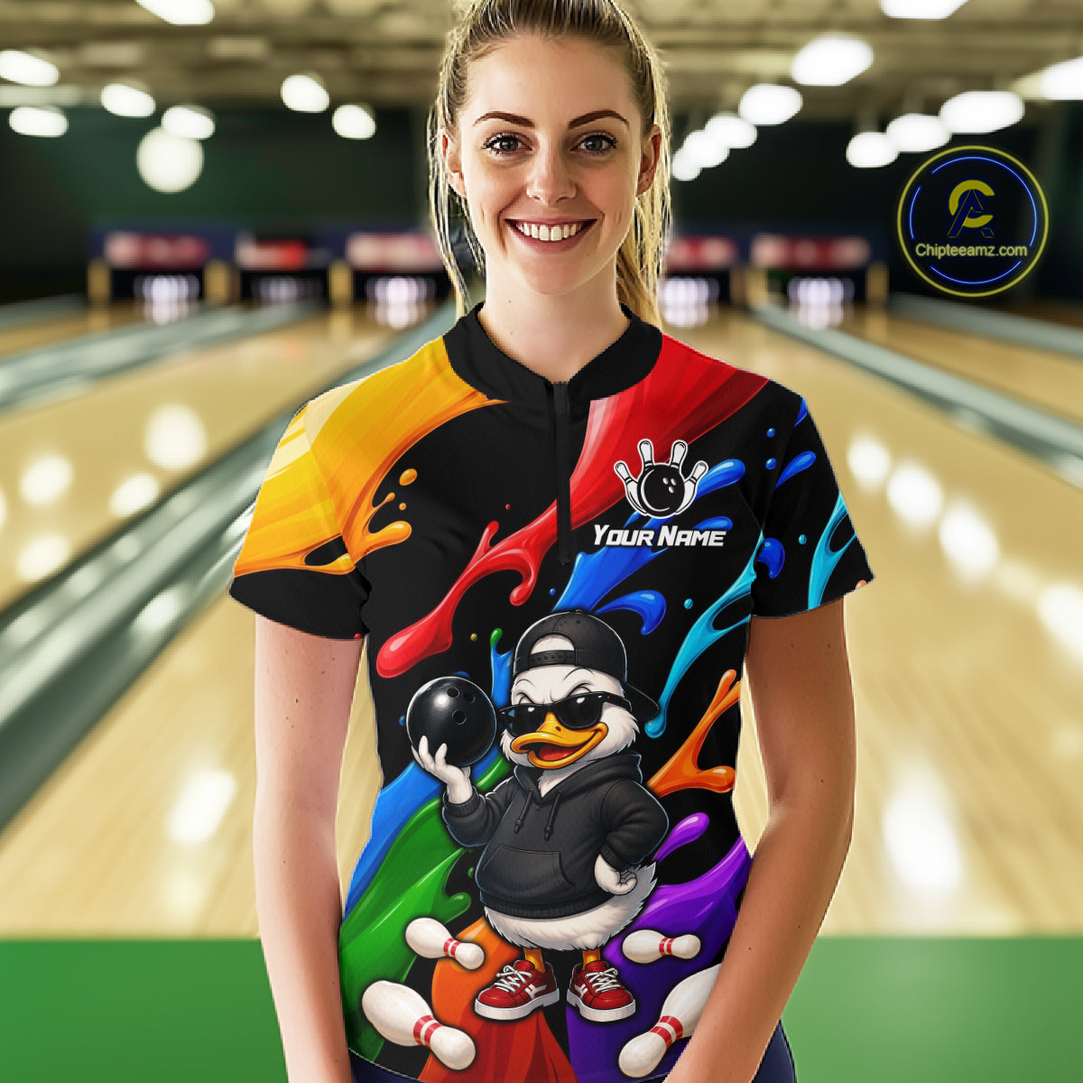 A woman stands in a bowling alley wearing a short-sleeve bowling shirt with a colorful abstract paint-splatter design and a cartoon duck bowling graphic featuring the text 'YOUR NAME' 