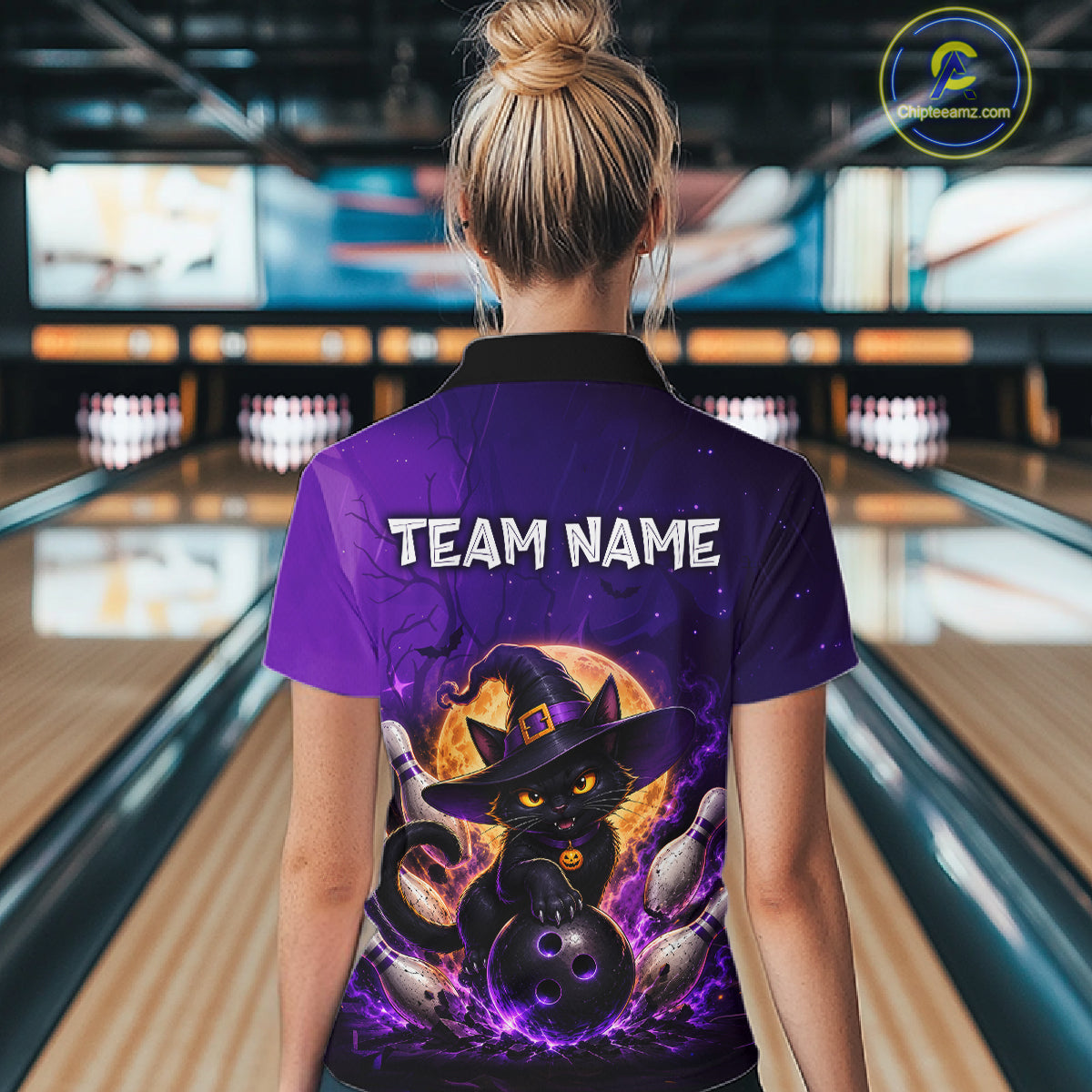 A woman viewed from behind wearing a purple bowling-themed polo shirt with a black cat in a witch hat graphic and the text 'TEAM NAME' on the back, standing in a bowling alley.