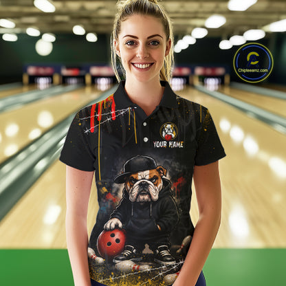 A woman stands in a bowling alley wearing a black short-sleeve polo shirt with a custom graphic print of a bulldog dressed as a detective holding a red bowling ball. The shirt features 'YOUR NAME' and a custom pin on the chest