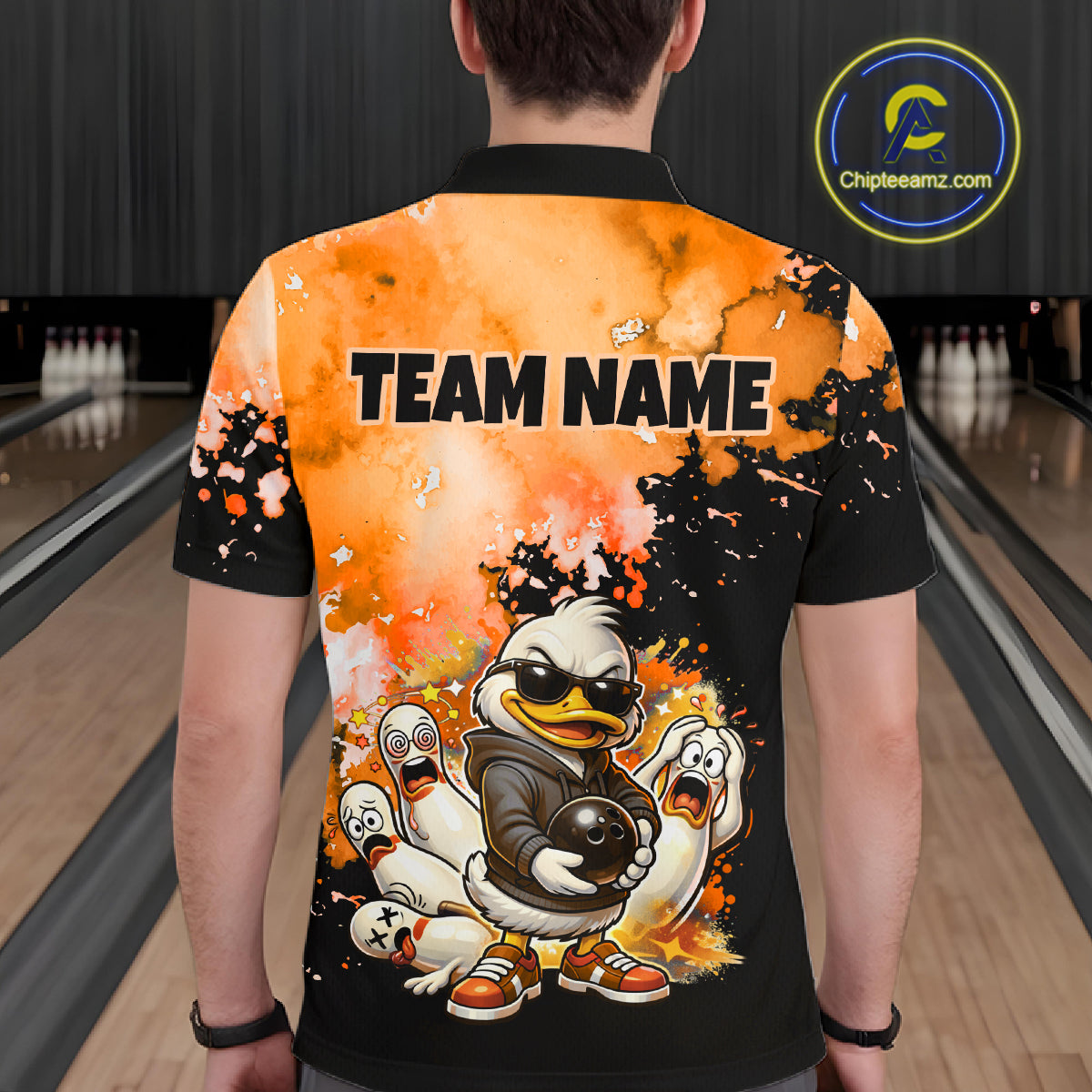 A person viewed from behind wearing a short-sleeve bowling shirt with a custom orange and black graphic design featuring cartoon ducks and the text 'TEAM NAME'. The background shows bowling lanes