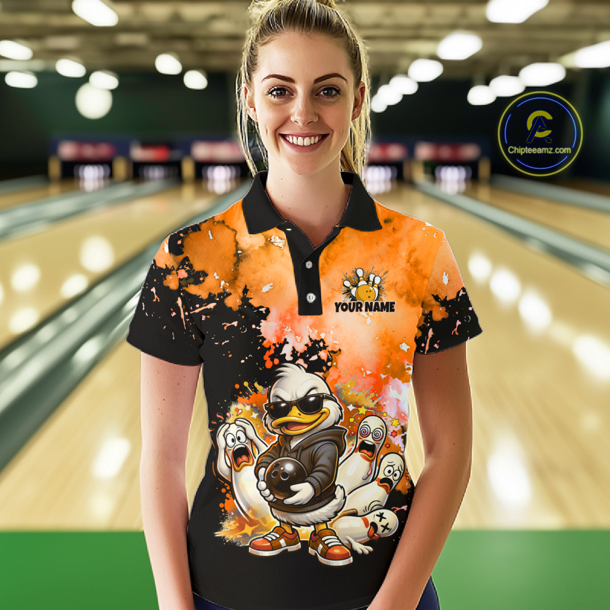 A woman stands in a bowling alley wearing a custom short-sleeve polo shirt with a cartoon duck graphic and 'YOUR NAME' text. The shirt features a black and orange splatter design 