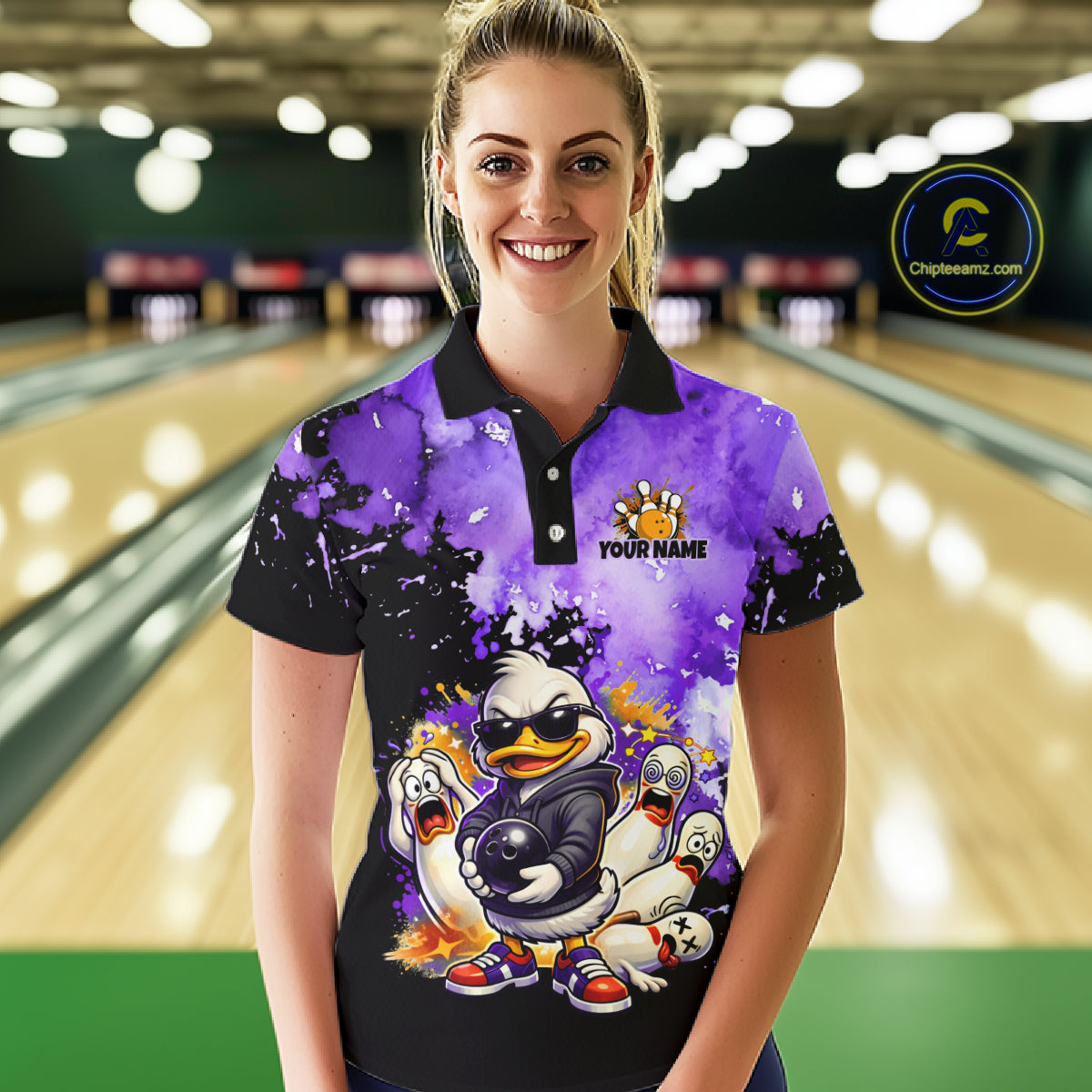 A woman stands in a bowling alley wearing a custom short-sleeve polo shirt with a purple and black splatter design and a cartoon duck character graphic. The shirt has a black collar and placket, with a purple and black splatter pattern and a cartoon duck graphic on the front.