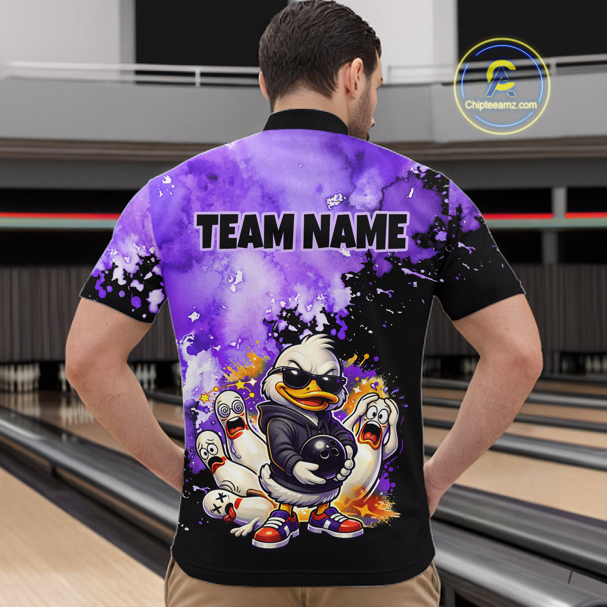 A man viewed from behind wearing a custom bowling-themed short-sleeve polo shirt with a purple and black watercolor design, cartoon ducks, and the text 'TEAM NAME' on the back. The background shows bowling lanes in a bowling alley.