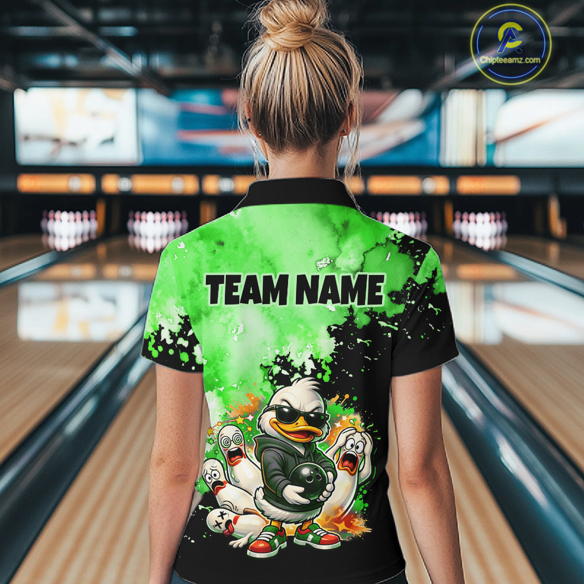 A woman viewed from behind wearing a short-sleeve collared bowling-themed shirt in a bowling alley. The back of the shirt is bright green and black with the text 'TEAM NAME' and a cartoon duck-and-feather bowling graphic.