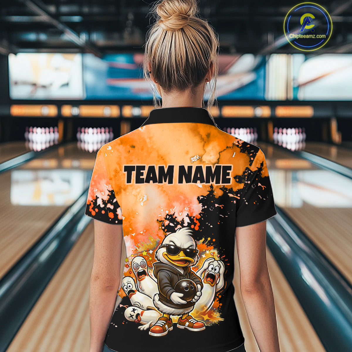 A woman viewed from behind wearing a short-sleeve collared bowling shirt with a fiery orange and black splatter design, cartoon ducks, and the text 'TEAM NAME' on the back. The background shows blurred bowling lanes in a bowling alley.