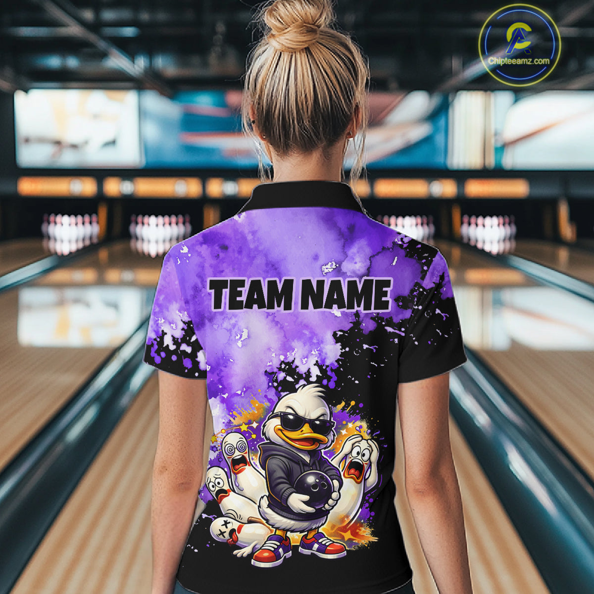 A woman viewed from behind wearing a custom short-sleeve polo shirt in a bowling alley. The back of the shirt features a purple and black watercolor design with a cartoon duck character, skulls, and the text 'TEAM NAME'.