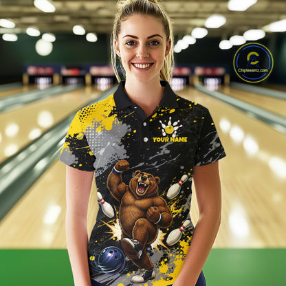 A woman stands in a bowling alley wearing a custom black polo shirt with a colorful graphic of a bear catching bowling pins, yellow paint splatters, and the text 'YOUR NAME'.
