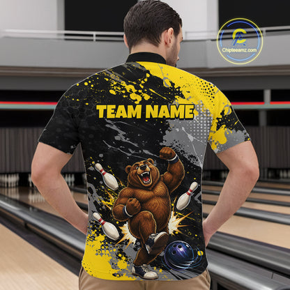 A man viewed from behind wearing a custom bowling-themed short-sleeve shirt with a graphic of a roaring brown bear bowling, yellow and black paint splatters, and the text 'TEAM NAME' on the back. The background shows a blurred bowling alley interior.