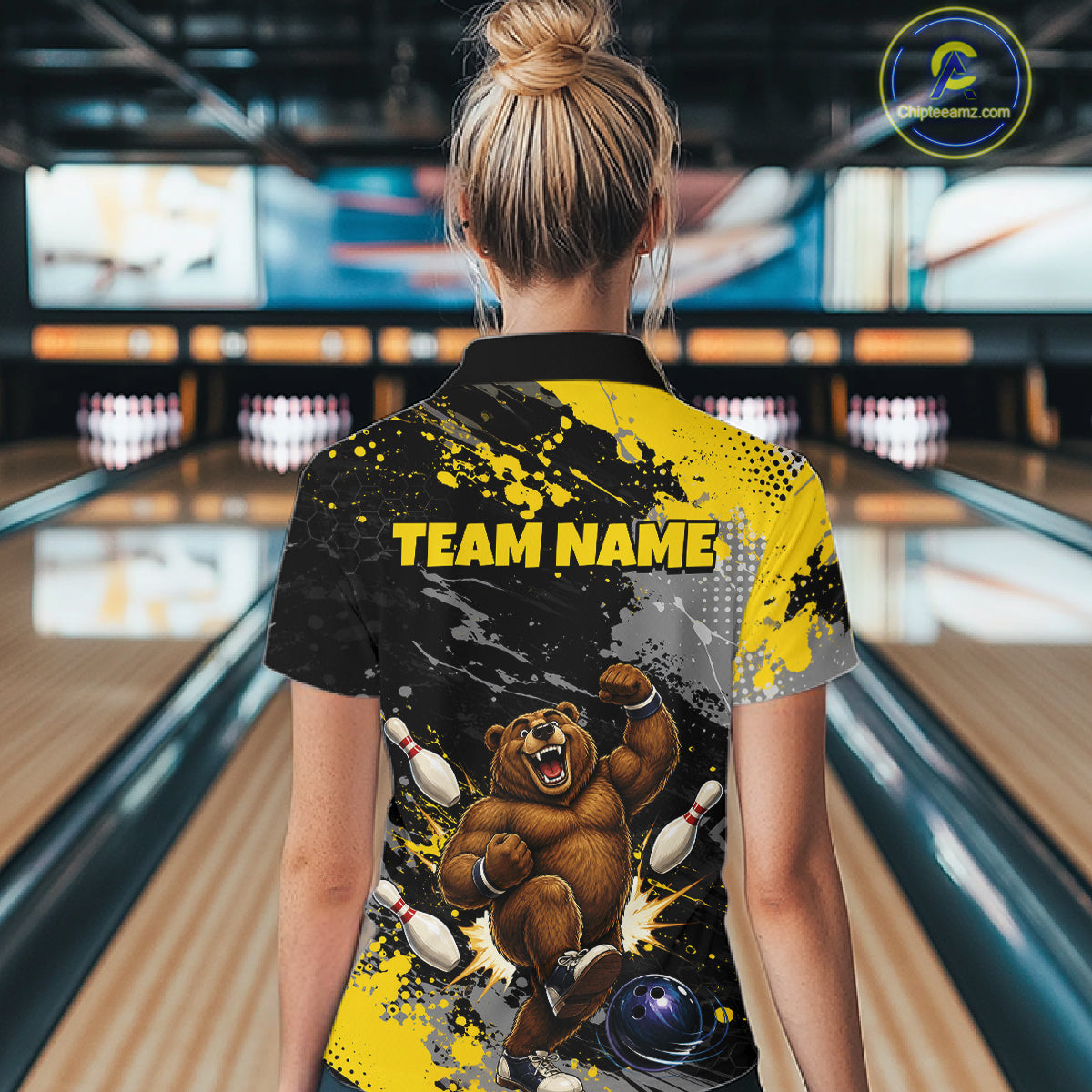 A woman viewed from behind wearing a custom bowling-themed short-sleeve shirt with a black and yellow paint splatter design, a bear graphic, and 'TEAM NAME' printed on the back, set against a bowling alley background.