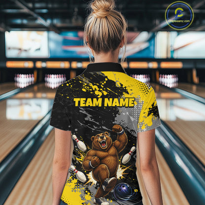 A woman viewed from behind wearing a custom bowling-themed short-sleeve shirt with a black and yellow paint splatter design, a bear graphic, and 'TEAM NAME' printed on the back, set against a bowling alley background.