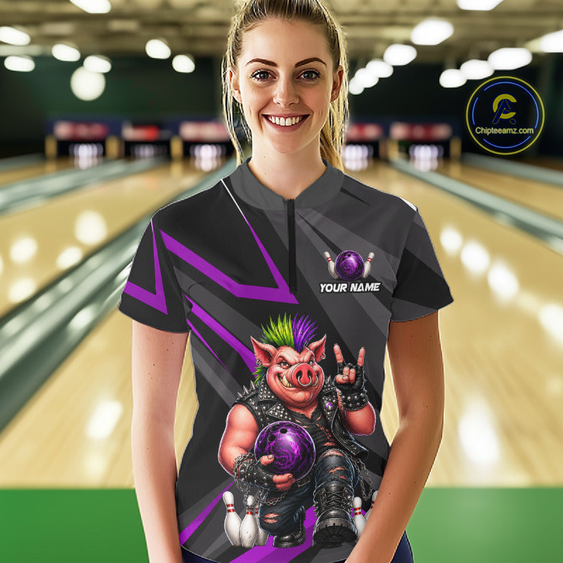 A woman stands in a bowling alley wearing a custom short-sleeve bowling jersey. The jersey is dark gray with purple graphic stripes and a large graphic of a pig character holding a purple bowling ball.