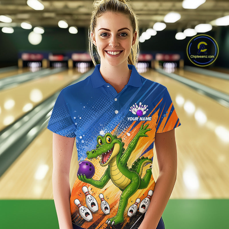 A woman stands in a bowling alley wearing a custom blue polo shirt with a colorful graphic of a green alligator holding a purple bowling ball, with bowling pins and orange streaks on the front. 