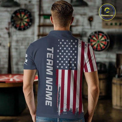 A man viewed from behind wearing a navy blue short-sleeve polo shirt with a distressed American flag print on the back and 'TEAM NAME' printed vertically in white. The background shows a blurred indoor bar or gym setting with dartboards.