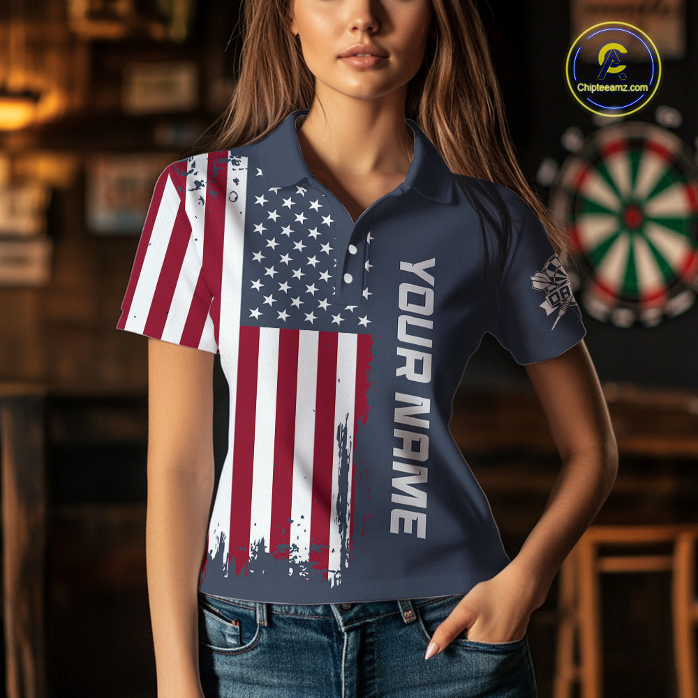 A woman wearing a navy blue custom polo shirt featuring a distressed American flag print on the right side and vertical white text reading 'YOUR NAME' on the left chest. The background is a dimly lit bar or lounge with a dartboard visible.
