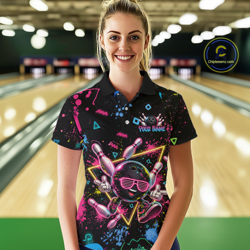 A woman stands in a bowling alley wearing a black short-sleeve polo shirt with a colorful retro bowling-themed graphic print featuring bowling pins, balls, and neon splatter details. The background shows blurred bowling lanes and lights.