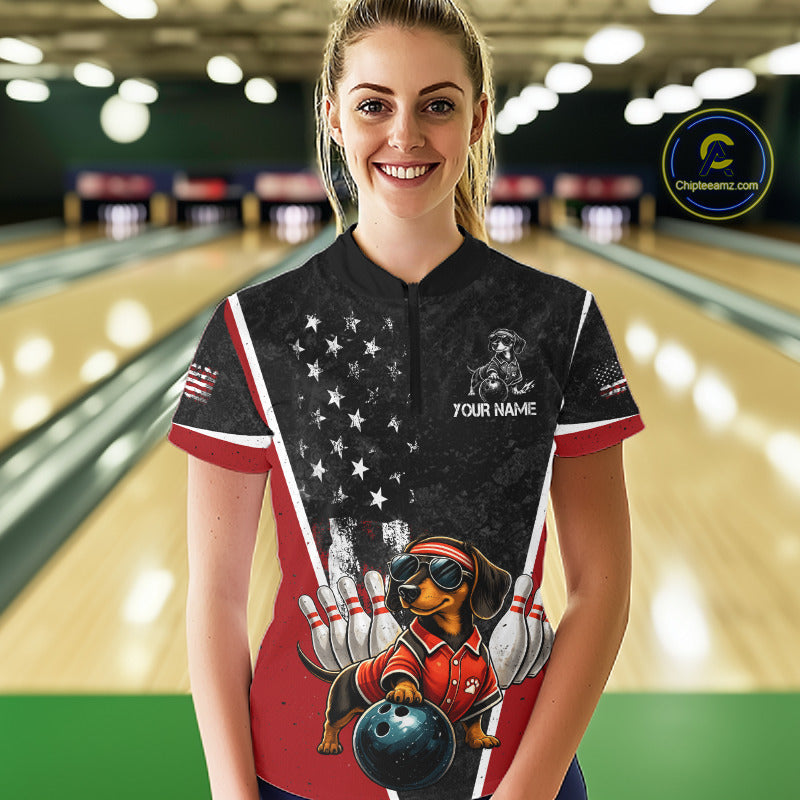 A woman stands in a bowling alley wearing a custom printed bowling-themed short-sleeve polo shirt. The shirt features a black upper section with a white star pattern resembling the American flag, red and white side stripes, a dachshund bowling graphic, and placeholder text 'YOUR NAME'.