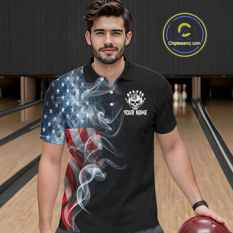 A man wearing a black short-sleeve polo shirt with a graphic design featuring the American flag and swirling smoke, standing in a bowling alley. The shirt has a white skull and crown logo on the left chest with the text 'YOUR NAME' below it.