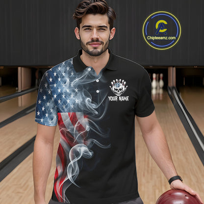 A man wearing a black short-sleeve polo shirt with a graphic design featuring the American flag and swirling smoke, standing in a bowling alley. The shirt has a white skull and crown logo on the left chest with the text 'YOUR NAME' below it.