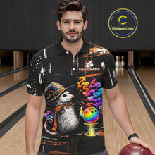 Load image into Gallery viewer, A man wearing a black short-sleeve bowling-themed graphic polo shirt with a colorful cartoon opossum, bowling ball, and spray-paint splatter design, standing in a bowling alley. 