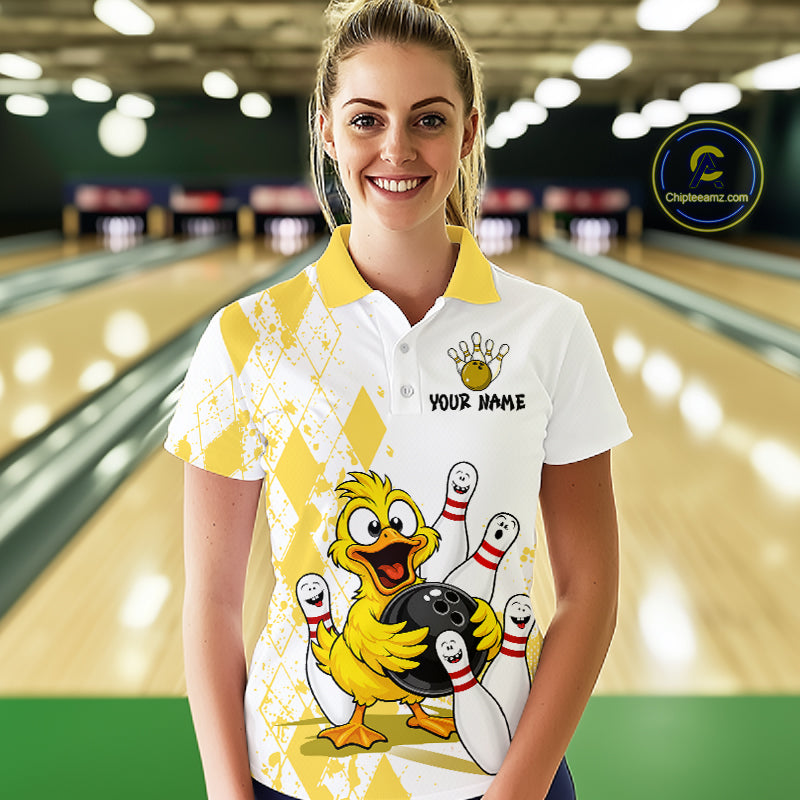 A woman stands in a bowling alley wearing a white and yellow bowling-themed polo shirt with a cartoon duck holding bowling pins and a bowling ball. The shirt has a yellow collar, a bowling pin graphic, and the text 'YOUR NAME' on the chest.