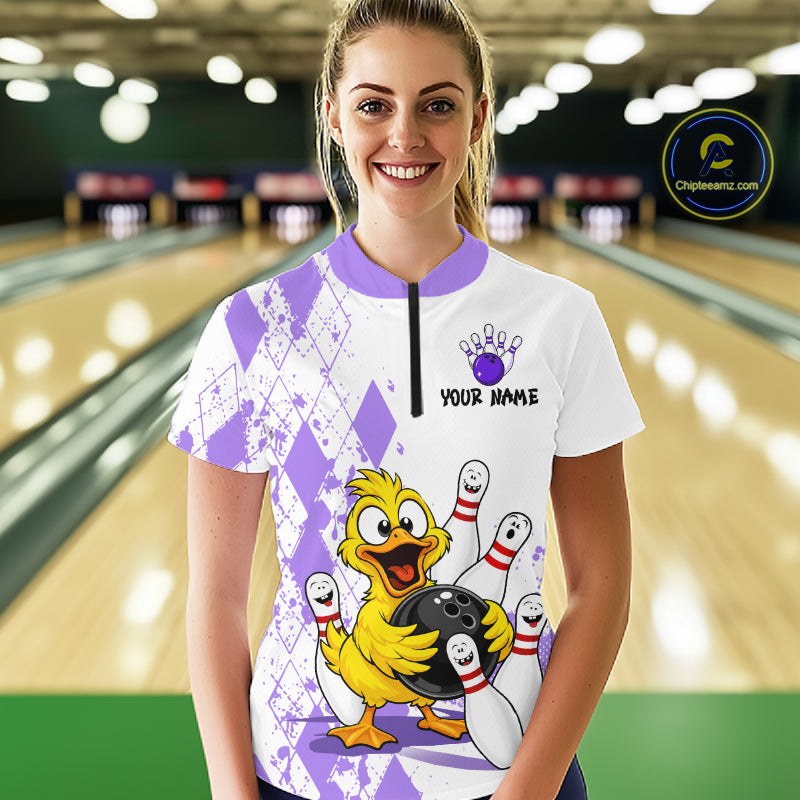A woman stands in a bowling alley wearing a white short-sleeve bowling-themed t-shirt with purple argyle patterns, a cartoon yellow duck holding a bowling ball, and a custom name print on the chest.
