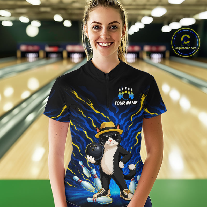 A woman stands in a bowling alley wearing a black short-sleeve bowling t-shirt with a colorful graphic of a cat bowling and the text 'YOUR NAME'. The background shows blurred bowling lanes.