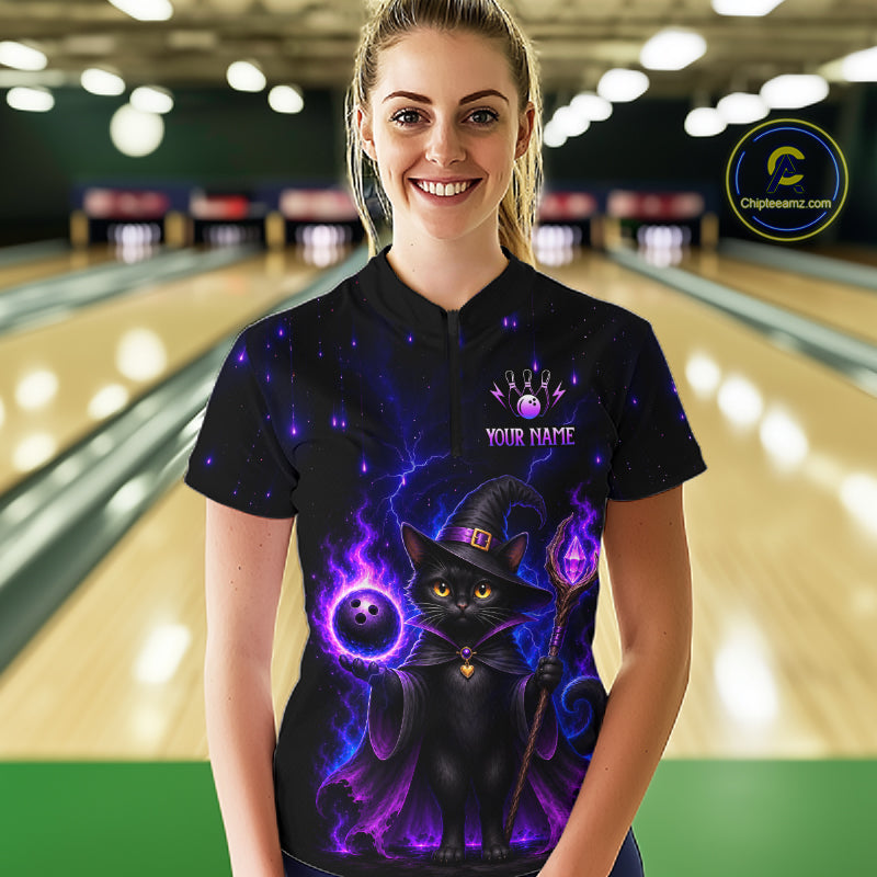 A woman stands in a bowling alley wearing a black short-sleeve polo shirt with a colorful graphic of a black cat dressed as a witch surrounded by purple magic, a bowling ball, and the text 'YOUR NAME'. 