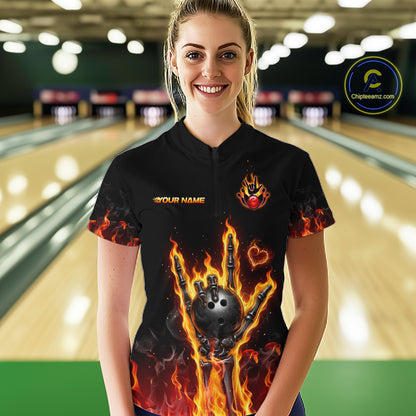 A woman stands in a bowling alley wearing a black short-sleeve bowling shirt with a fiery graphic of a bowling ball and flames, along with 'YOUR NAME' and a logo on the chest. 