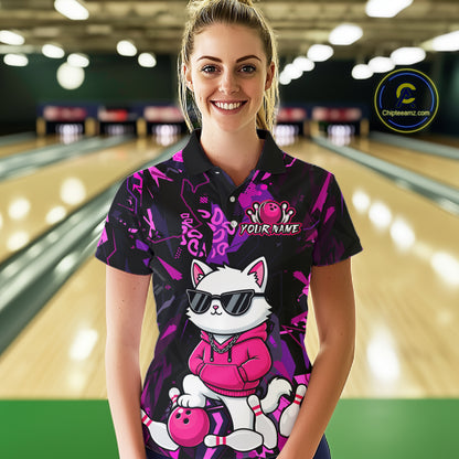 A woman standing in a bowling alley wearing a custom short-sleeve polo shirt with a purple and black background, featuring a cartoon white cat wearing sunglasses and a pink hoodie holding a bowling ball. The shirt has 'YOUR NAME' printed on the chest.