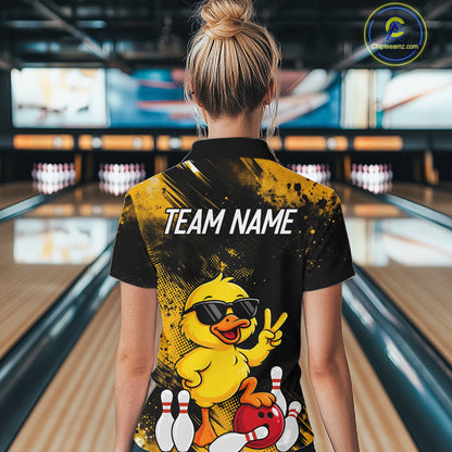 A woman viewed from behind wearing a black and yellow bowling-themed polo shirt with a cartoon duck graphic and the text 'TEAM NAME' on the back, standing in a bowling alley.